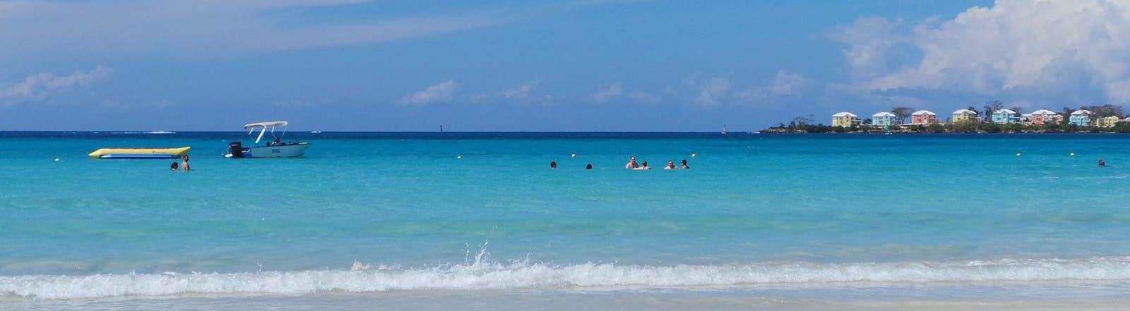 A beach at Negril, Jamaica in bright midday sun: broad pale sand, a breaking surf line, wide turquoise water with swimmers and a small boat, and a low green headland with pastel buildings on the horizon