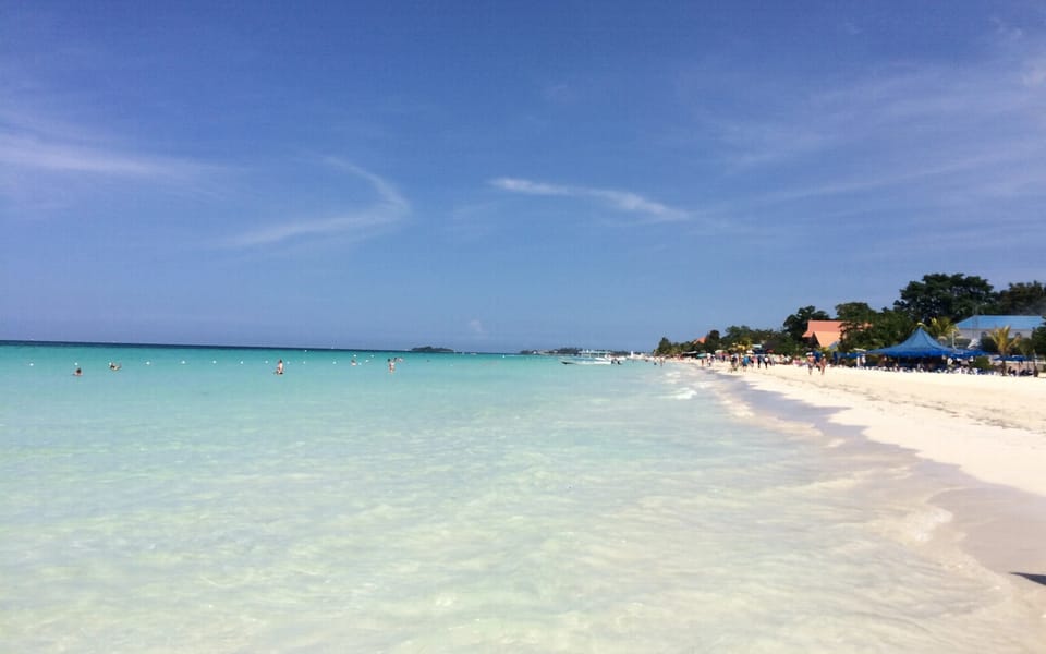 A long stretch of Negril beach in bright sunshine: clear shallow turquoise water in the foreground, white sand curving away to the right and low trees, buildings and blue beach shades in the distance.