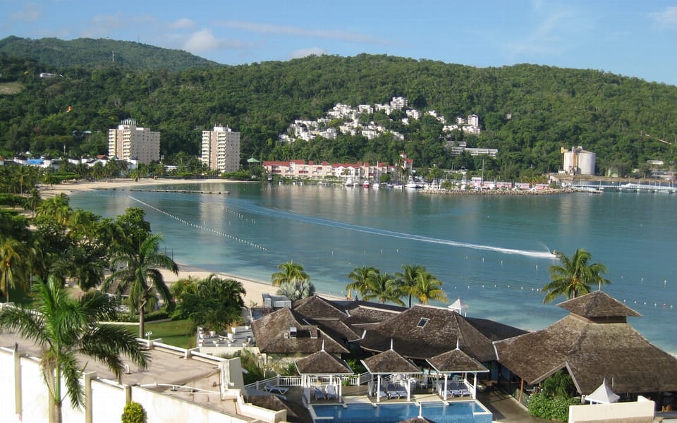 Ocho Rios harbour seen from above: a sheltered blue bay with moored boats and a marked swimming area, a curving sand beach, palms and low resort roofs in the foreground and a green hillside covered in white buildings behind.