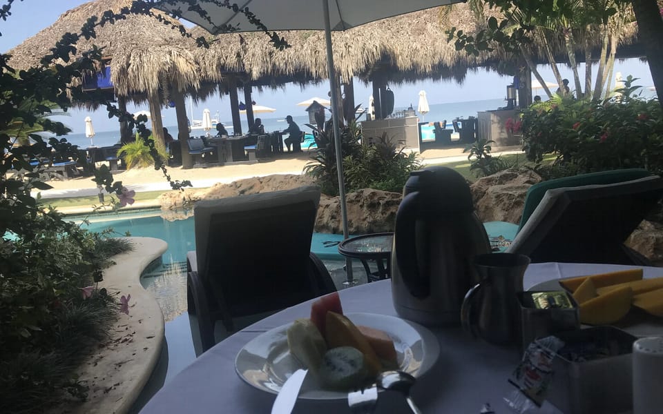 A turquoise swim-up pool with a curved stone edge and flowering shrubs, with a thatched-roof beach bar, pale sand and the calm sea beyond it.