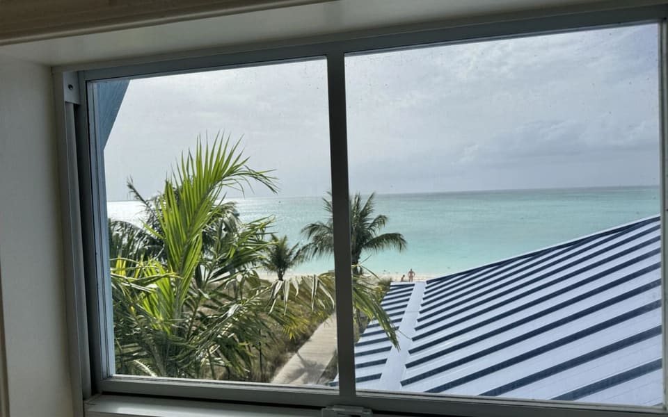 The beach and turquoise water seen through a sliding window from inside the villa, with palms and a striped roof in the foreground.