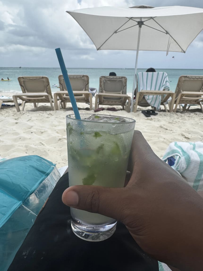 A held glass of a mint and lime drink in the foreground on the beach, looking past empty timber loungers and a white parasol to clear turquoise water under a light overcast sky.