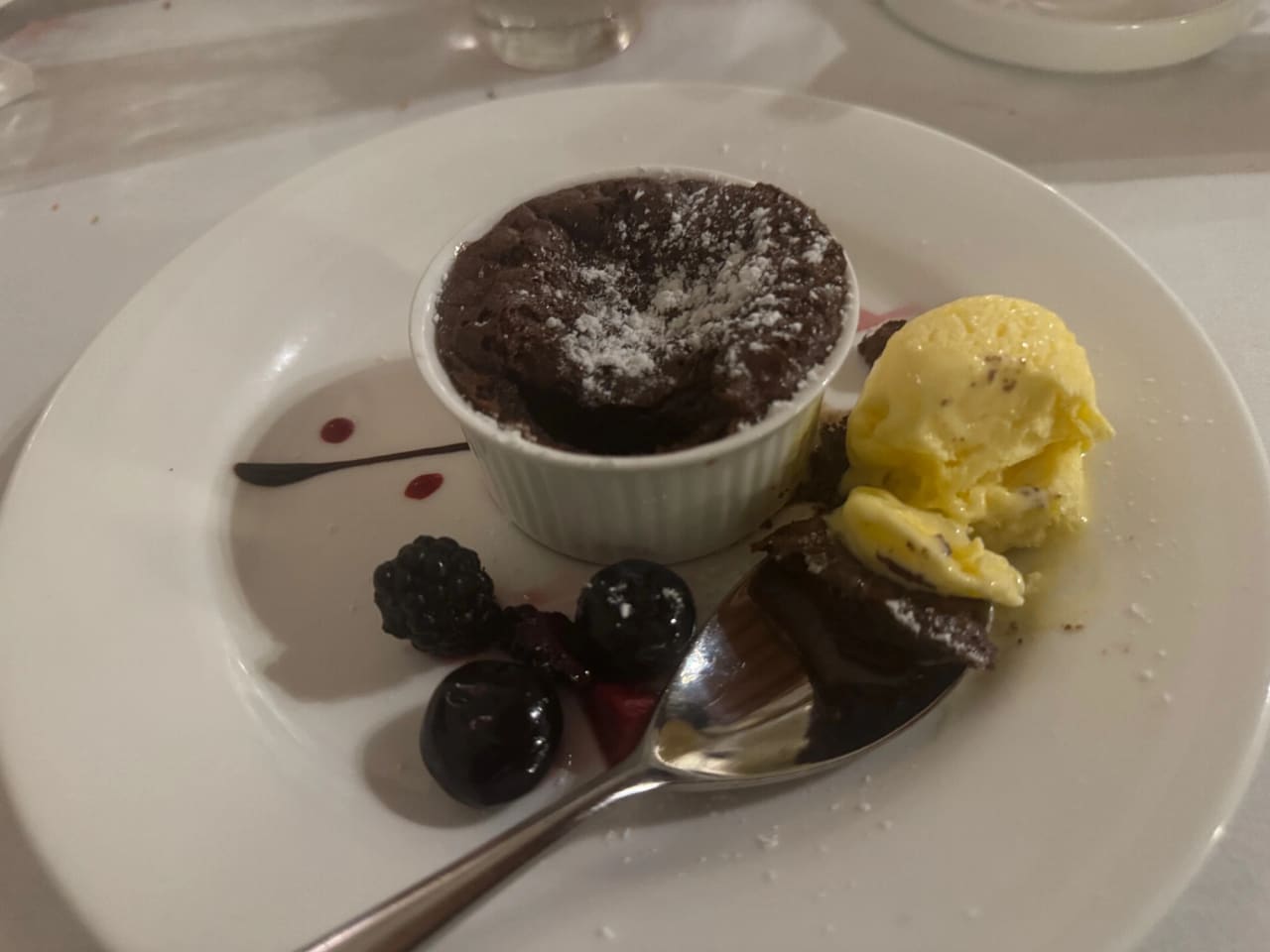 A dessert plate holding a chocolate pudding in a white ramekin dusted with icing sugar, a scoop of vanilla ice cream, a slice of chocolate cake, blackberries, blueberries and a cherry, with chocolate and berry sauce drizzled across the plate.