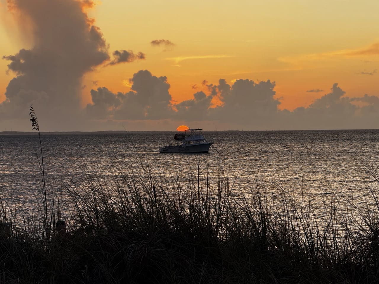 Sunset over calm water photographed through silhouetted sea grass, with an orange sky, a tall bank of cloud, the sun low on the horizon and a moored cabin boat in the middle of the frame.
