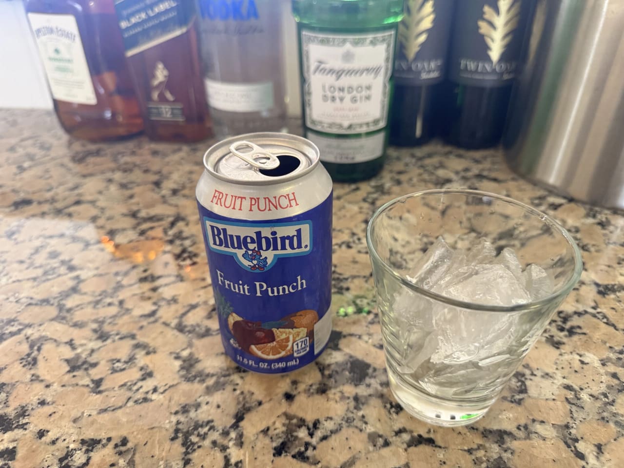 A granite kitchen counter in the villa holding an opened can of fruit punch and a glass of ice, with a row of spirit bottles and a metal cocktail shaker standing behind them.