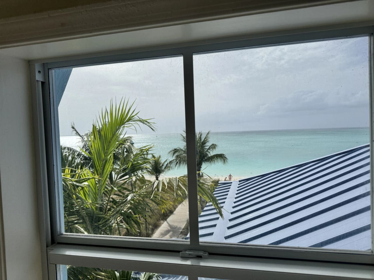 The beach seen through a sliding window from indoors: a white window frame around palms and low planting, a timber boardwalk running toward the sand, a blue-and-white striped roof in the near foreground, and flat turquoise water meeting a pale sky.