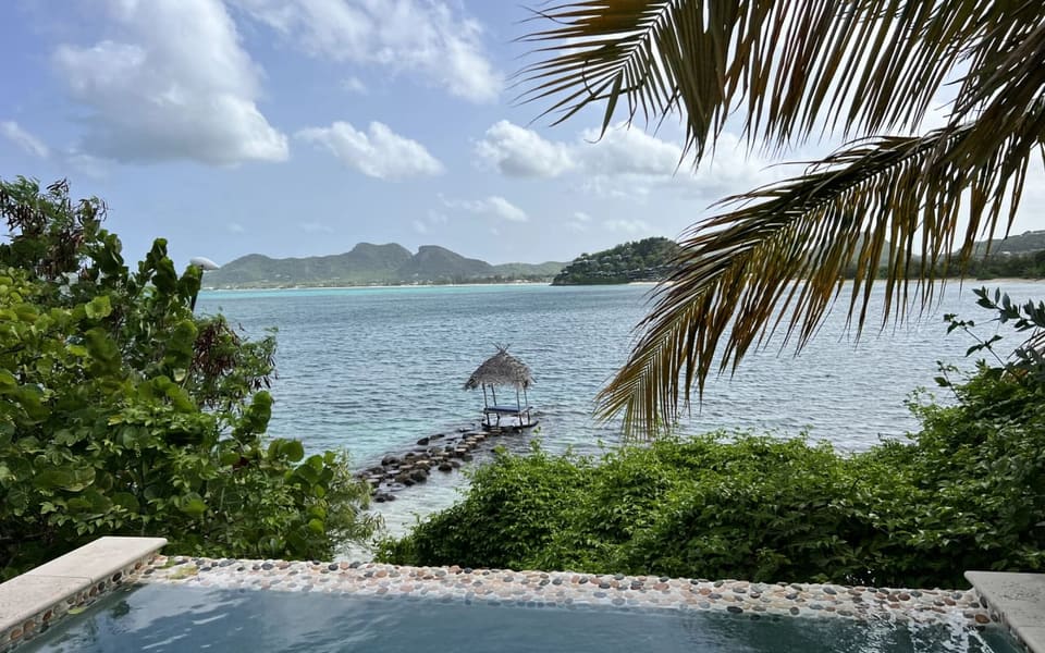 A tiled plunge pool in the foreground with a palm frond overhanging, and a turquoise bay with a thatched shade hut on a rock jetty beyond it.
