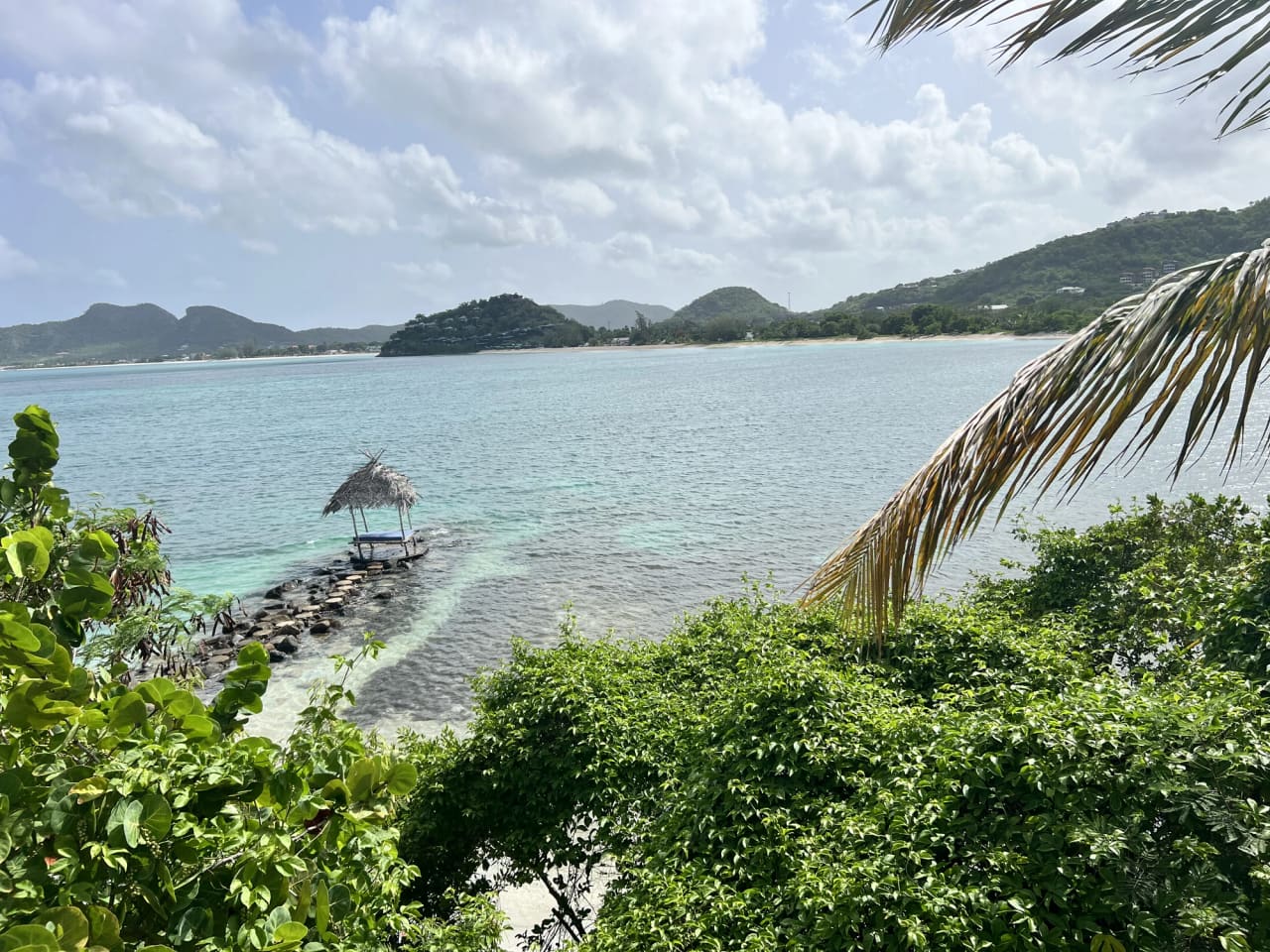 A bay seen from higher up the hillside: turquoise shallows shading to deeper blue, a thatched shade hut on a rock jetty at the water's edge, a narrow strip of beach, and green hills rising along the opposite shore under a bright sky.