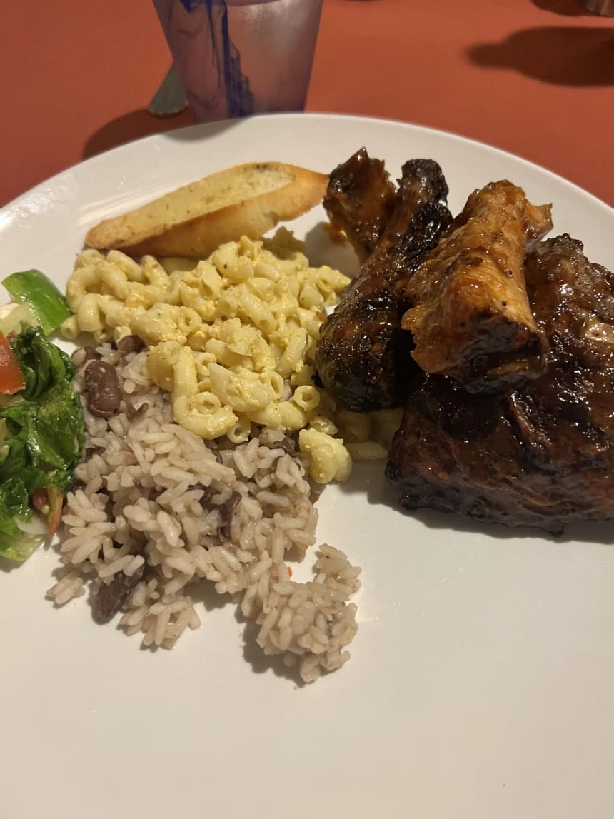 A dinner plate holding glazed barbecue chicken pieces, rice and peas, macaroni and cheese, a slice of garlic bread and a small side salad, photographed indoors in warm evening light.