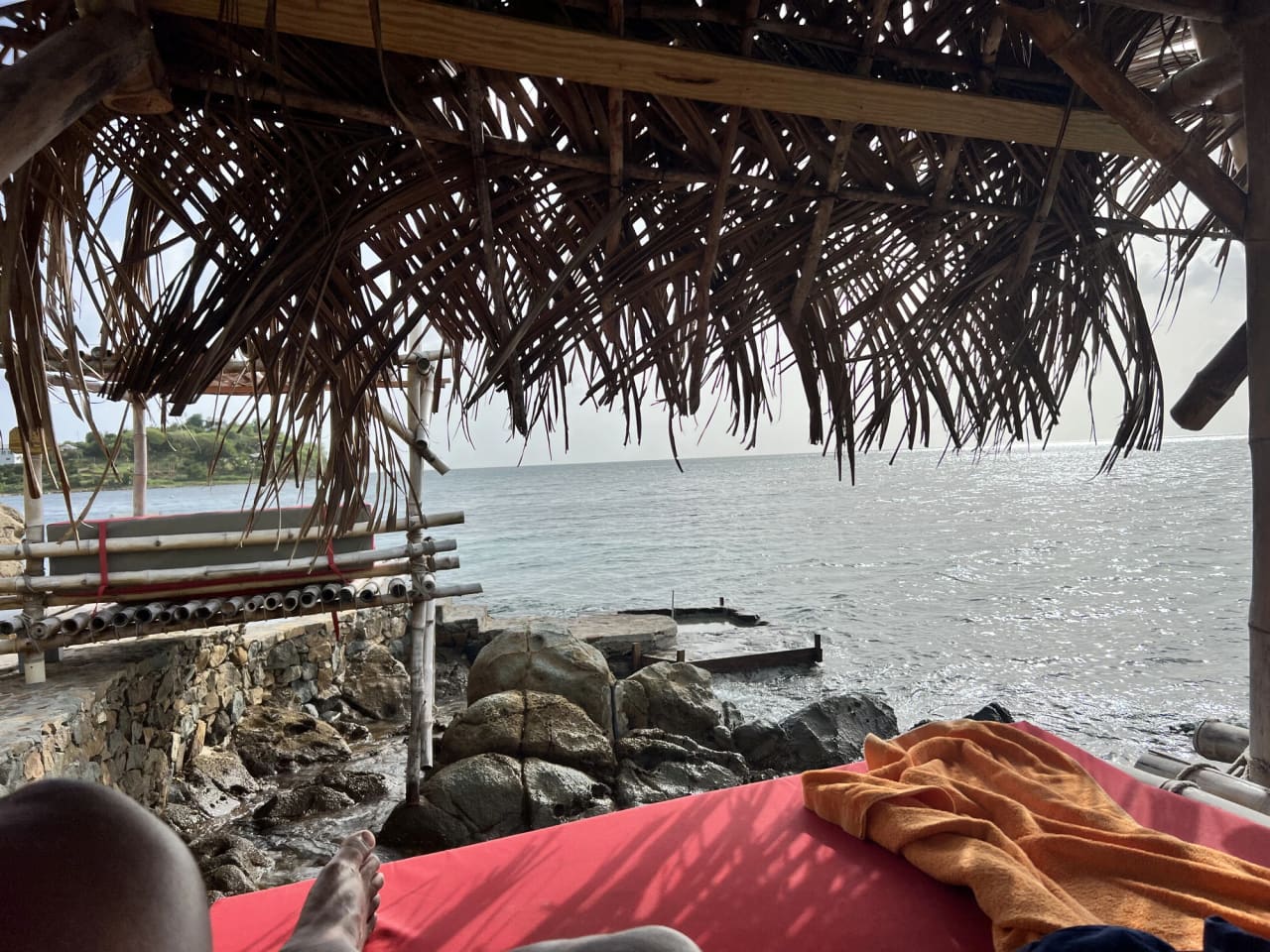 The view out to sea from a lounger under a palm-thatch shade hut. A red lounger cushion and an orange towel fill the near foreground, the rocky shoreline and a low rock platform sit at the water's edge, and a second thatched hut stands to the left with the far shore beyond.