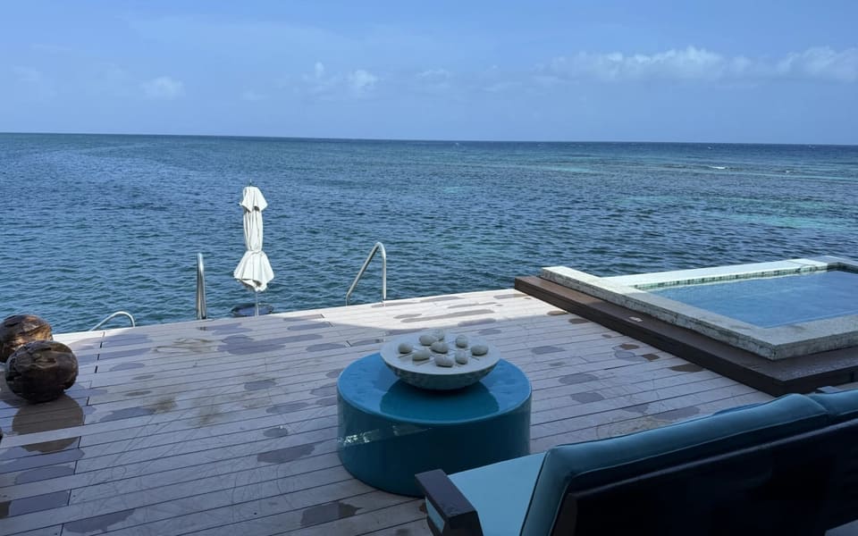 A wooden deck built out over the sea, with a low blue table, a plunge pool set flush with the deck and pale reef showing through the water.