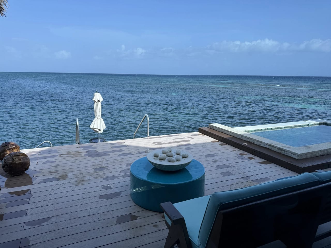 A wooden deck built out over the sea in bright daylight. A low round blue table holds a stone bowl set out as noughts and crosses, husked coconuts sit on the deck boards at the left, a tiled plunge pool sits flush with the deck on the right, a furled white parasol stands in the water beside a steel handrail, and pale reef shows through the water toward the horizon.