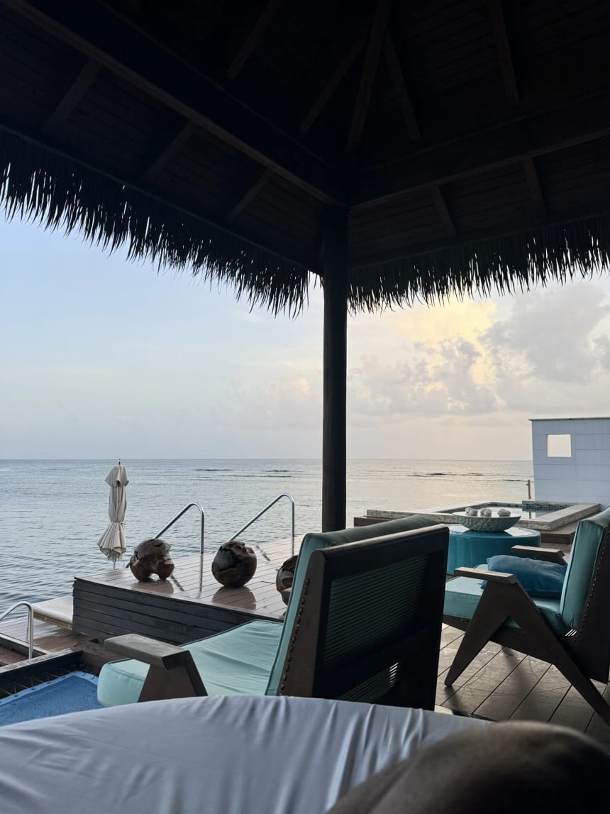 Looking out from the day bed under a thatched roof across the covered part of the overwater deck in soft early light. A pale cushion and a shadowed knee are in the foreground, dark timber chairs with teal cushions and a low table sit to the right beside the plunge pool, husked coconuts rest on the deck boards, and a furled parasol stands in flat calm water beside a steel handrail under a pale sky.