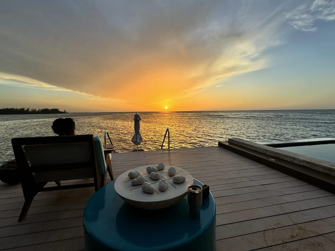 The overwater deck at sunset. The sun sits just above the horizon, throwing an orange path across the water, with a low wooded point silhouetted at the far left. Someone sits in a low timber chair at the left with their back to the camera, looking out at it, and the blue table with the stone noughts-and-crosses bowl is in the foreground.
