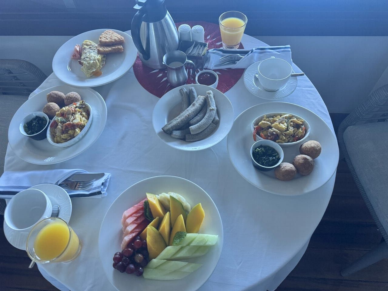 A breakfast for two seen from above on a round white-clothed table: two plates of ackee and saltfish with fried dumplings and small dishes of dark green callaloo, a plate of sliced mango, melon, watermelon and grapes, a dish of boiled green bananas, a covered plate, two glasses of orange juice, cups and a steel coffee pot.