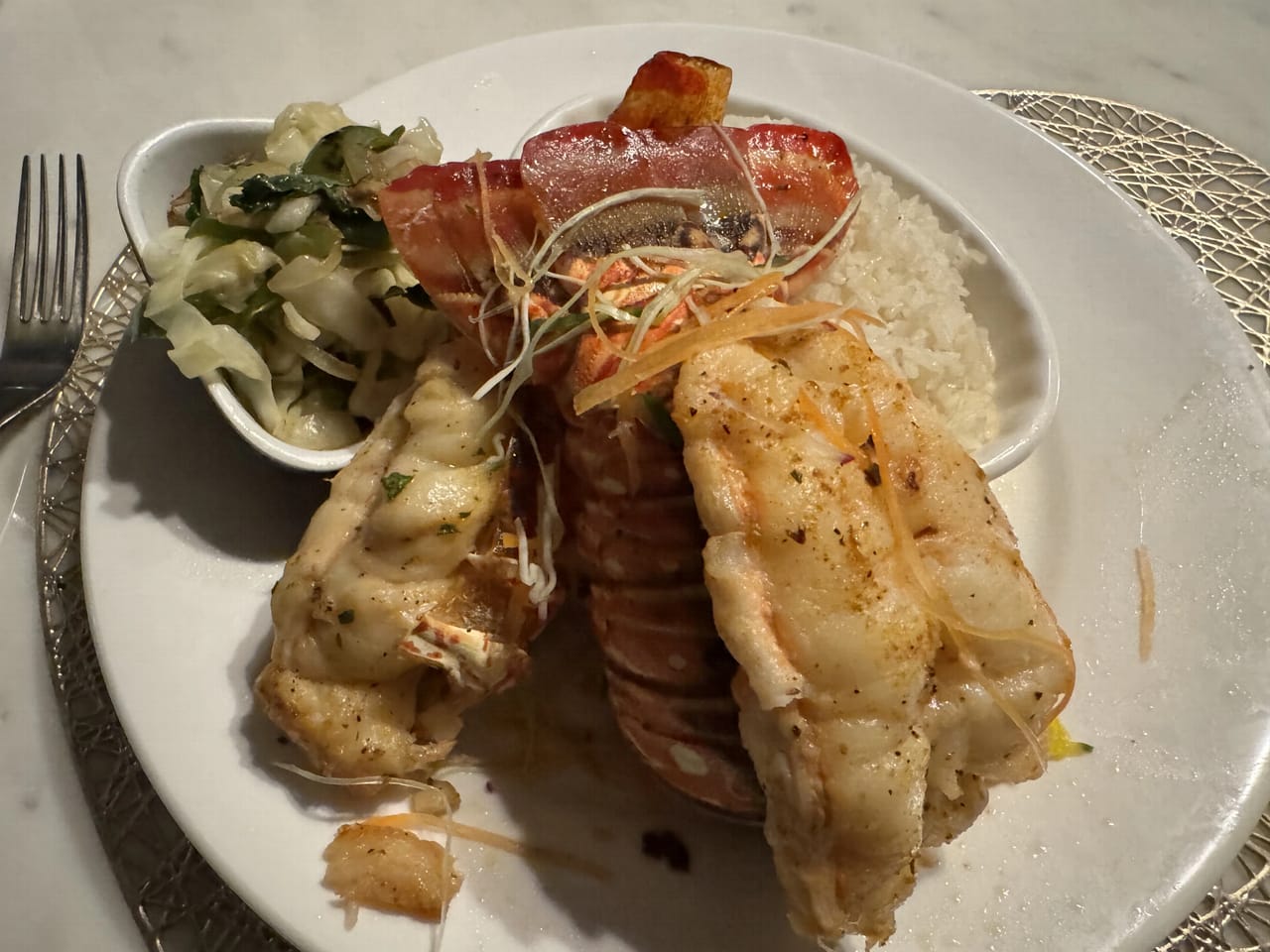 A dinner plate seen from above under warm indoor light: two grilled lobster tails out of the shell laid across the plate, a bacon-wrapped portion behind them, a small bowl of cabbage slaw and a fork to the left, and white rice to the right.