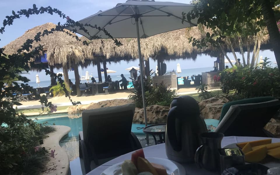 A turquoise swim-up pool with a curved stone edge and flowering shrubs, with a strip of sand, a thatched-roof beach bar and the calm sea beyond it.