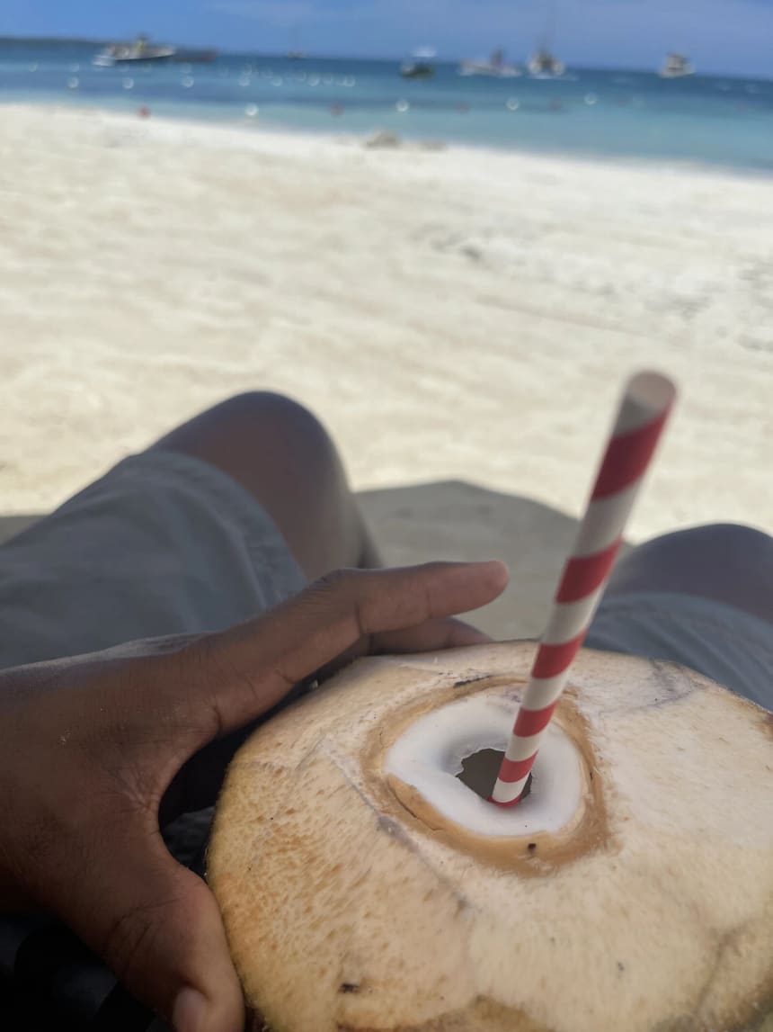 A hand holding a fresh young coconut with its husk trimmed flat and a red and white striped paper straw pushed into the opening, with a soft-focus white sand beach, turquoise sea and small boats behind.