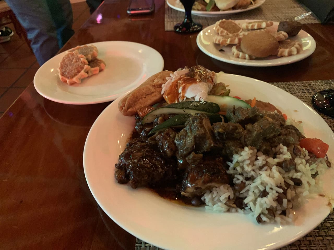 A plate of curry goat with rice and peas, a bread roll, sliced cucumber and a piece of fish, on a polished dark wooden table, with side plates of fritters and festival behind it.