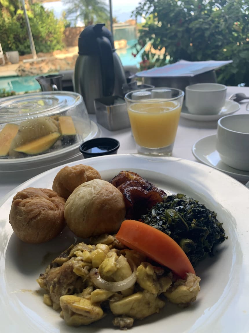 A plate of ackee and saltfish with fried dumplings, dark green callaloo, a wedge of papaya and fried plantain, on an outdoor table with a glass of orange juice, an insulated coffee flask, white cups and a covered dish of sliced fruit behind it.