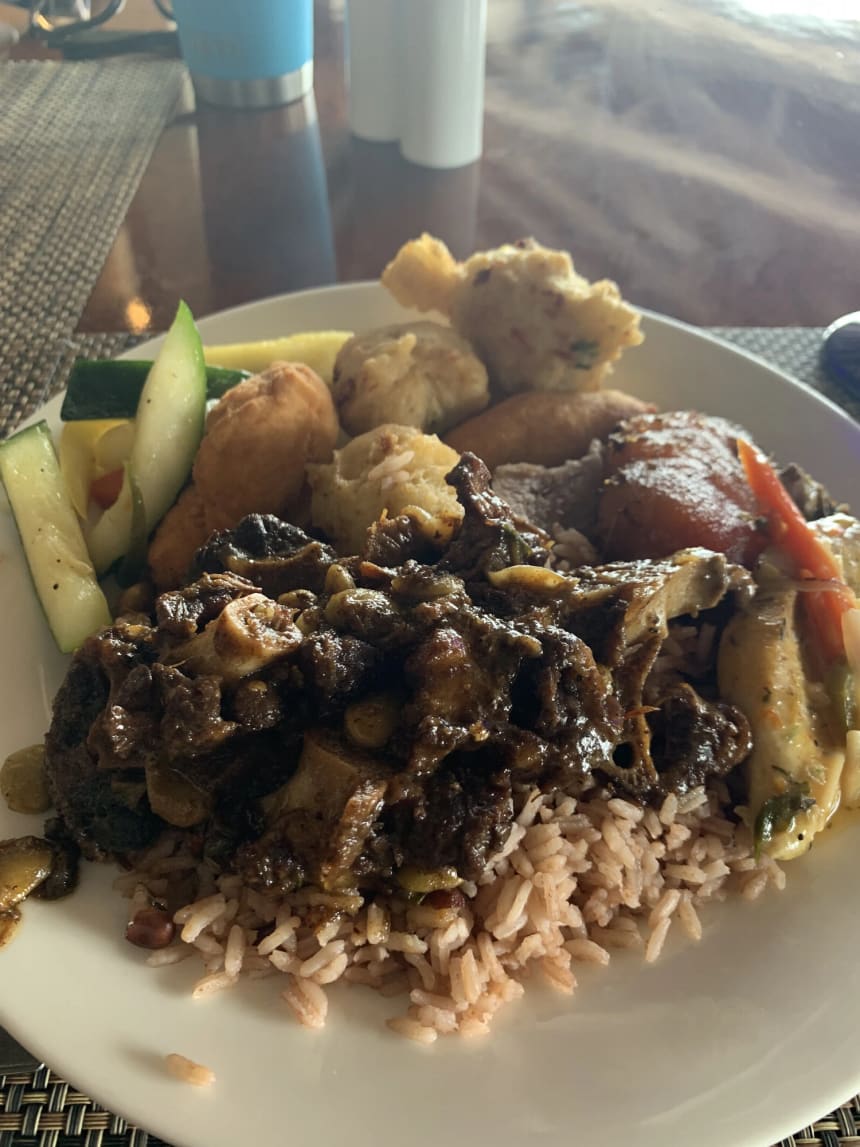 A plate of oxtail in dark gravy served over rice and peas, with festival and fish fritters, fried plantain, sliced cucumber and squash, on a woven placemat.