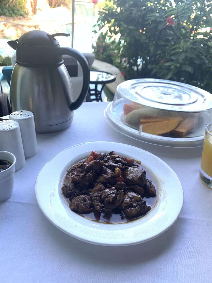 A plate of Jamaican stew chicken in dark gravy on a white plate at an outdoor table, with a steel coffee flask, salt and pepper shakers, a glass of juice and a covered dish of sliced papaya and melon beside it.